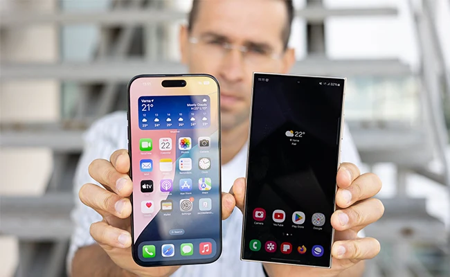 مقایسه دوربین جلوی یا سلفی آیفون ۱۶ با S24 Ultra تصویری از نمای جلوی گوشی S24 Ultra و گوشی iphone 16 pro max در دستان چپ و راست یک مرد